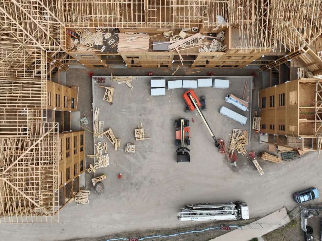 Top-down drone view of an active construction site courtyard with telehandlers, building materials, and wood-framed apartment buildings