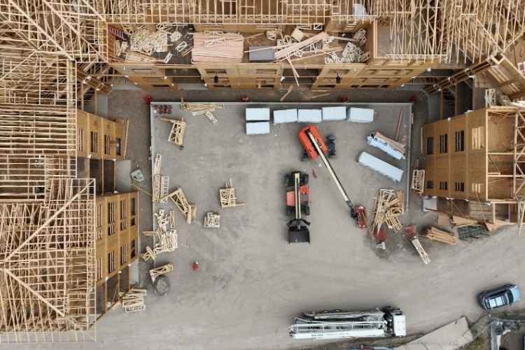 Top-down drone view of an active construction site courtyard with telehandlers, building materials, and wood-framed apartment buildings